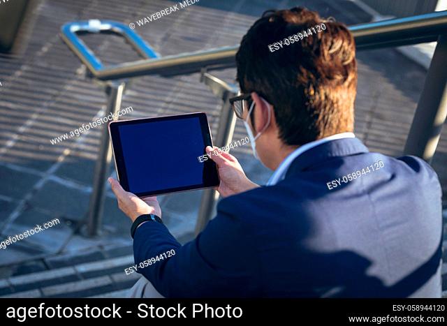 Asian businessman wearing face mask using tablet sitting on steps in city street