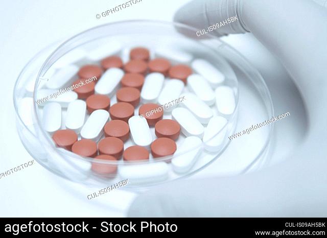 Hand holding a cover over a Petri dish with generic pills forming dollar symbol