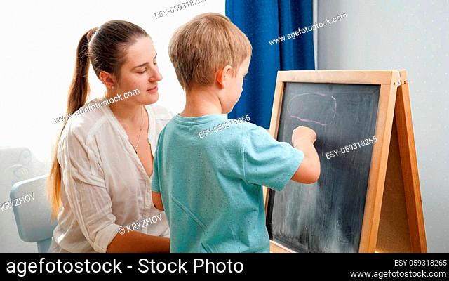 Little smiling boy learning ABC with young mother at home. Teacher writing alphabet on blackboard with chalk. Home education and remote school