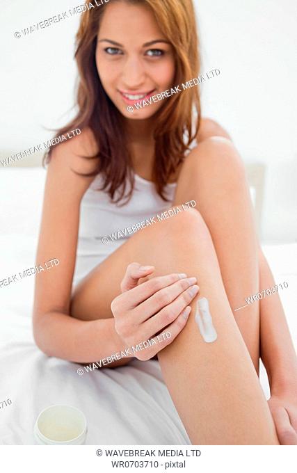 Portrait of a smiling young woman applying moisturizer on her leg while sitting on her bed