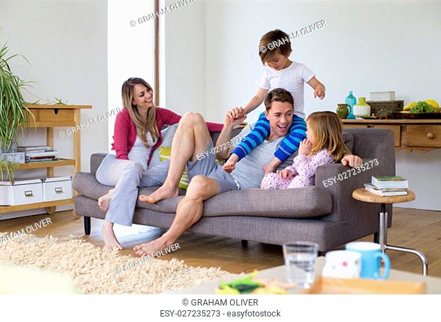 Family of four are sitting on the sofa in the living room of their home together. The father has his son on his shoulders and the little girl is sitting next to...
