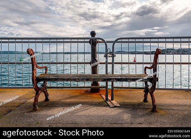A damaged bench without backrest and a barrier in front of it, seen in Torquay, Torbay, England, UK