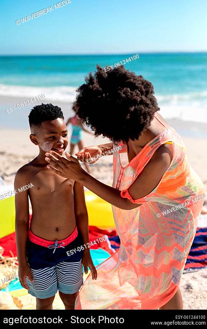 African american mother applying suntan lotion on son's cheek while enjoying summer holiday at beach