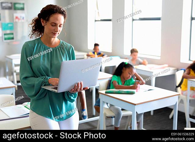 Caucasian young female teacher using laptop with multiracial elementary students in background