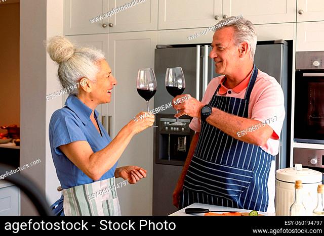 Happy caucasian senior couple standing in kitchen, drinking wine and preparing meal together