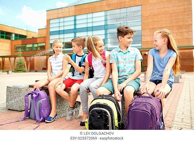 primary education, friendship, childhood, communication and people concept - group of happy elementary school students with backpacks sitting on bench and...