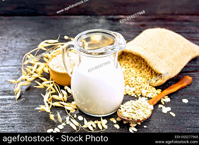 Oat milk in a jug, flour in bowl, oatmeal in a spoon, grain in bag, oaten stalks on the background of dark wooden board