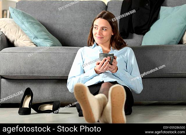 Satisfied businesswoman resting after work sitting on the floor with slippers drinking coffee at home
