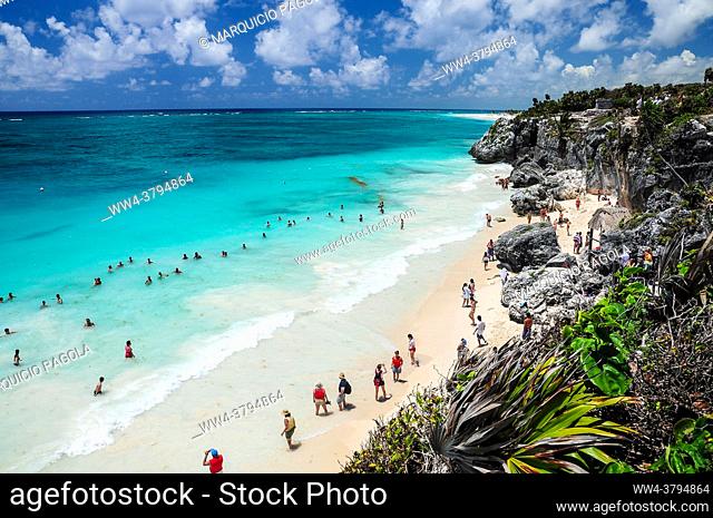 Ocean and rocky coast with lots of tourists walking on the beach in Tulum, Quintana Roo, Mexico. Some mayan ruins can be seen on the background