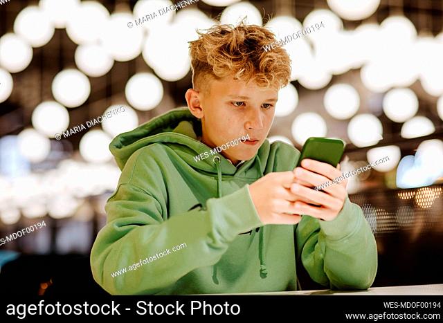 Teenage boy using smart phone in front of lights at food court