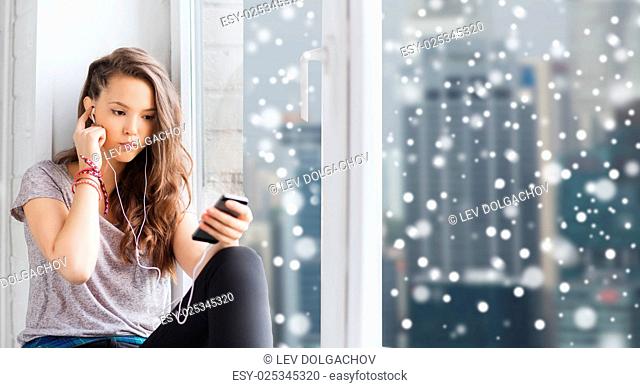 people, technology, winter, christmas and teens concept - sad pretty teenage girl sitting at window with smartphone and earphones listening to music over city...
