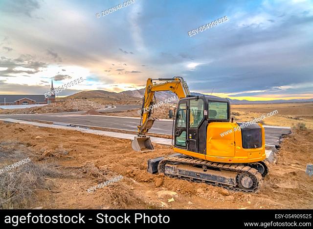 Excavator in Utah construction area near church. Excavator with grader blade beside a winding road in Utah Valley. A church, mountain, cloudy sky