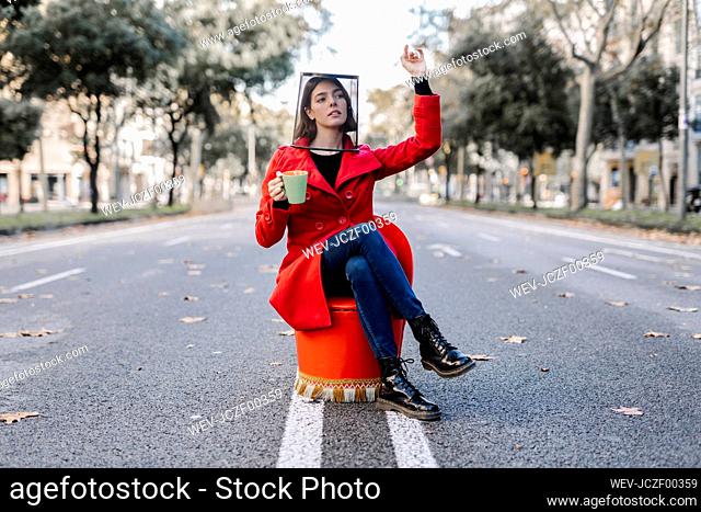 Young woman with coffee cup wearing garbage bin on head while sitting on chair at street