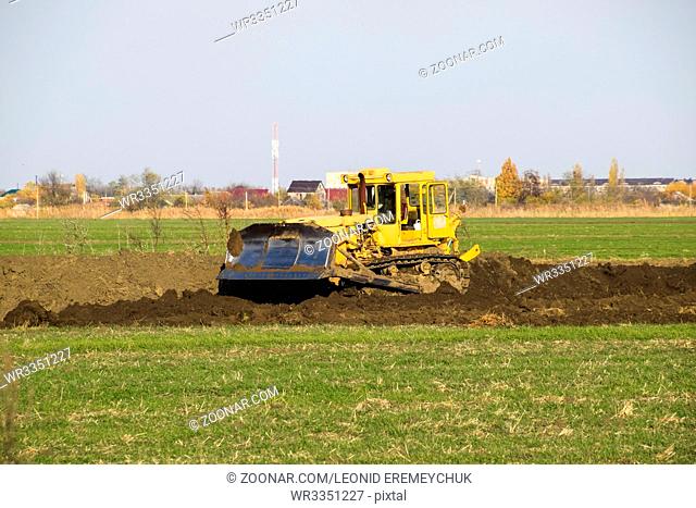 The yellow tractor with attached grederom makes ground leveling. Work on the drainage system in the field