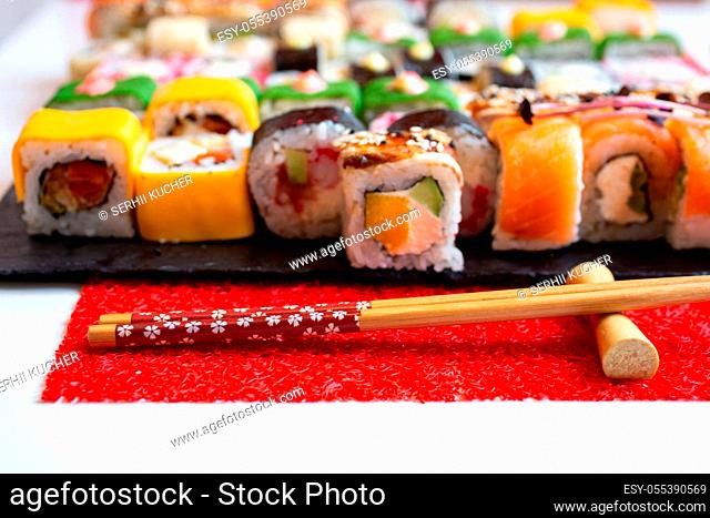 Wooden chopsticks on a red mat in the foreground. In the background, different colored sushi rolls on a stone black sushi board. Blurred background