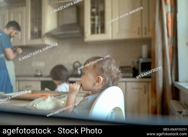 Baby boy eating pizza dough in kitchen seen through window