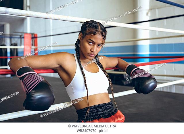 Female boxer with boxing gloves leaning on ropes and looking at camera in boxing ring. Strong female fighter in boxing gym training hard