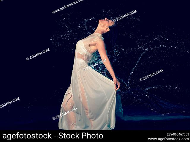 beautiful woman of Caucasian appearance with black hair dances in drops of water on a black background. The woman is wearing a white chiffon dress
