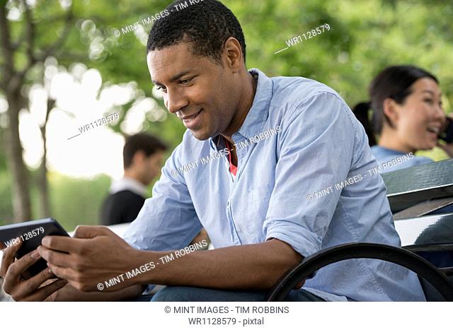 Summer in the city. People outdoors, keeping in touch while on the move. A man sitting on a bench using a digital tablet