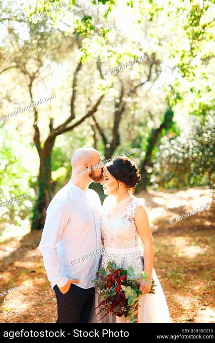 The bride and groom with a bouquet stand hugging among the trees in an olive grove, the groom kisses the bride on the forehead . High quality photo