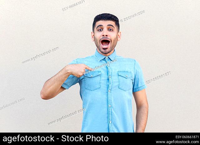 Portrait of surprised handsome young bearded man in blue shirt standing, pointing himself and looking at camera with amazed face