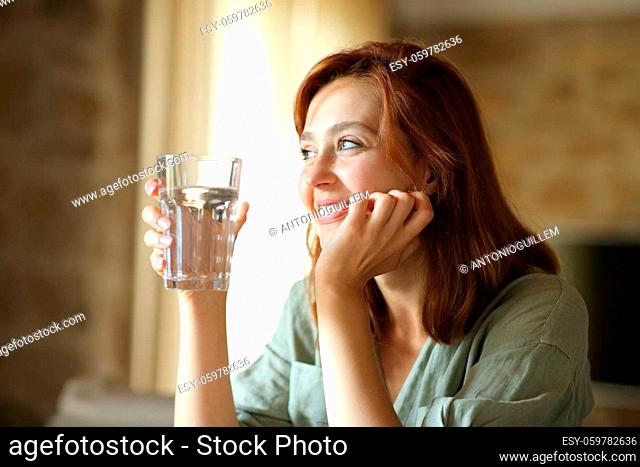 Happy woman holding water glass sitting in the living room at home looking away