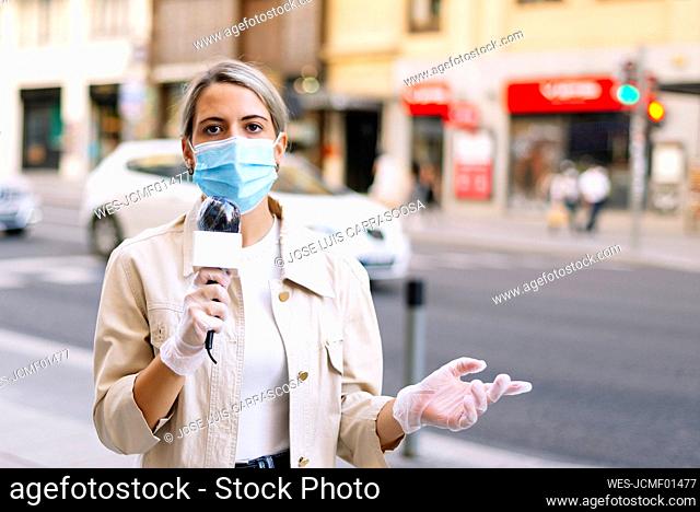 Female reporter wearing mask talking over microphone while standing on street in city