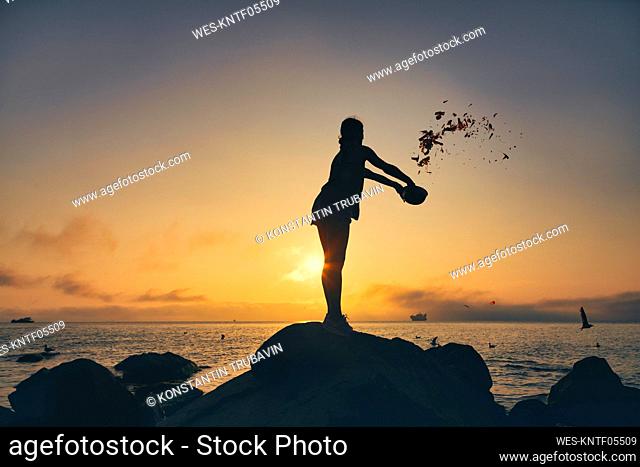 Silhouette woman feeding birds while throwing food from bowl at beach during sunset