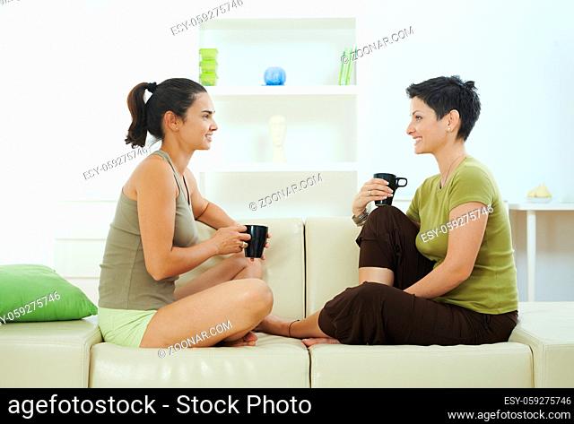 Two sisters sitting together on couch at home, drining coffee, smiling