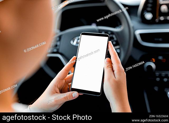 Mobile phone mockup with isolated white screen in the hand of a young woman. Interior view of modern SUV car. Female model with smartphone in the car