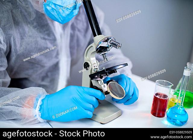 Medical professional in white protective suit looking through microscope in chemical laboratory. Search vaccine coronavirus. Covid 19 drug inventions