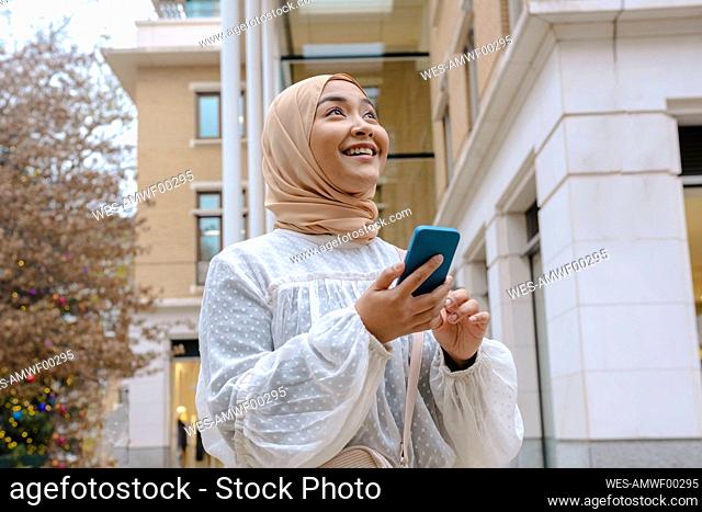 Happy young woman with smart phone standing in front of building