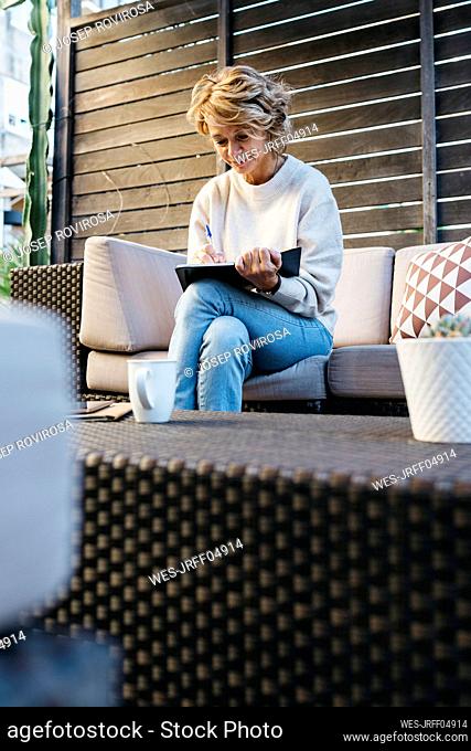 Mature woman writing in diary sitting on sofa at rooftop garden
