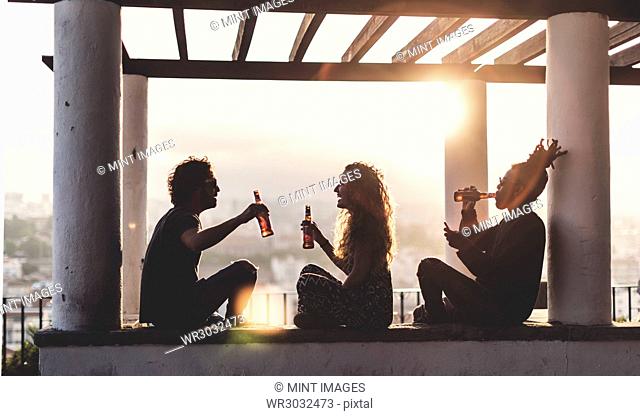 Silhouette of two men and a woman sitting outdoors under a pergola at sunset, holding beer bottles