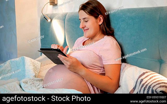 Portrait of smiling pregnant woman with big belly lying in bed and using tablet computer at night before going to sleep