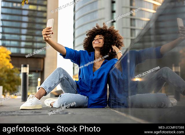 Smiling Afro woman taking selfie through mobile phone leaning on wall