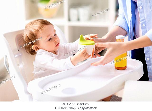 family, eating, feeding and people concept - mother giving spout cup with juice to baby at home
