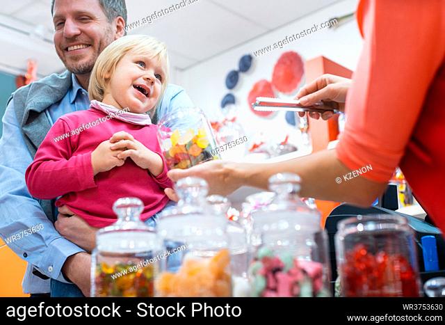 Saleslady for sweet things handing some candy to a kid being held by her father