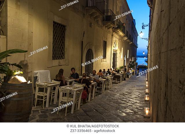 Tables and chairs are set up in the street at dusk outside a popular restaurant, Ortigia, Siracusa, Sicily. The ancient stones, historic buildings