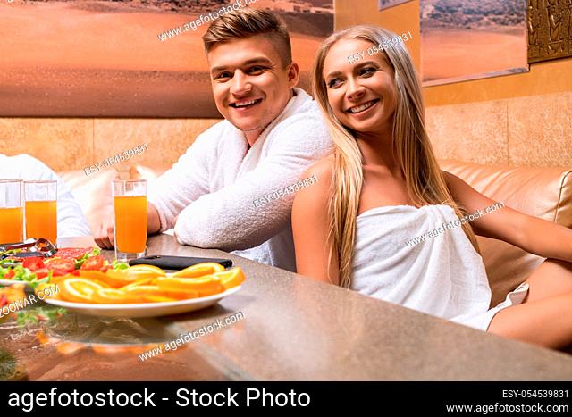 Smiling girl wrapped in a towel and her handsome boy friend wearing white bathrobe at table spending time in sauna