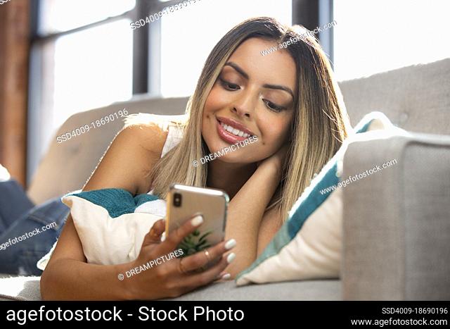 Young happy Hispanic woman using smart phone while lying on sofa at home