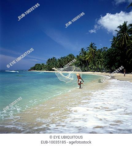 Fisherman casting fishing net into sea from the surf on a coconut palm tree lined sandy beach