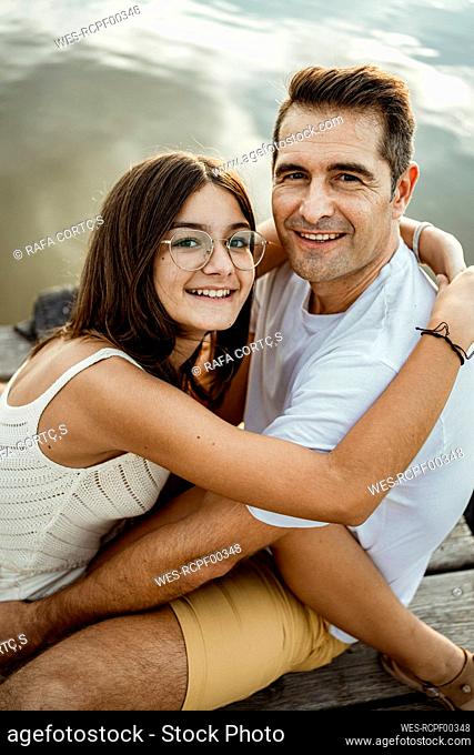 Smiling daughter embracing father while sitting at jetty