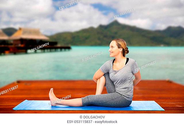 fitness, sport, people and healthy lifestyle concept - woman making yoga in twist pose on wooden pier over island beach and bungalow background