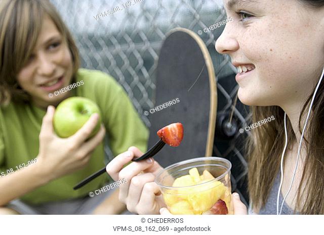 Close-up of a teenage boy holding an apple and looking at a teenage girl eating fruit salad