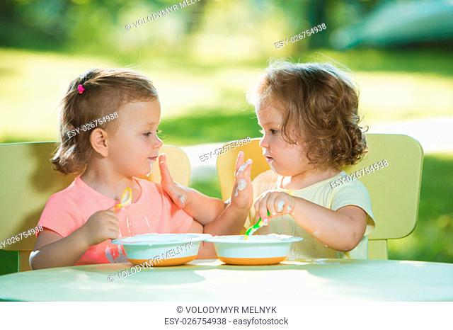 Two little 2 years old girls sitting at a table and eating together against a green lawn