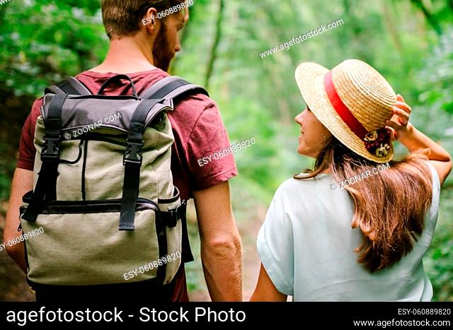 Guy and girl walk together in wood along the trail, holding hands. Back view. Happy couple holding hands and walking forest path