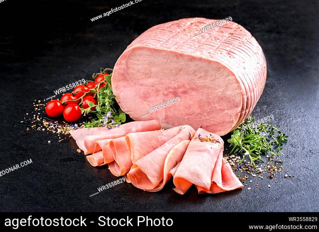 Traditional German boiled ham as piece and sliced decorated with tomatoes and herbs as top view on black slate slab with copy space