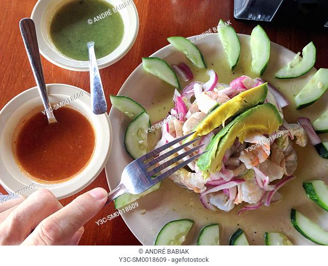 Male hand with fork above shrimp ceviche dish garnished with avocado wedges and cucumber pieces. Served with red and green salsa