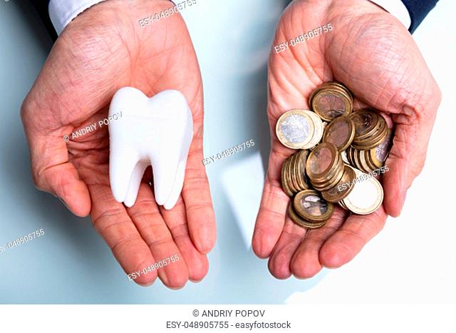 An Elevated View Of Man's Hand Holding White Tooth And Golden Coins Over Desk